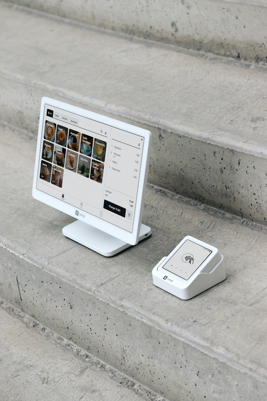 A point of sale (POS) setup is displayed on concrete steps, featuring a modern white display screen alongside a compact card reader. The screen shows a digital menu with various products, while the card reader is ready for contactless payments. This clean, minimalistic setup highlights the portability and sleek design of SumUp’s payment solutions, ideal for small businesses, cafes, and retail environments that value both functionality and aesthetics.